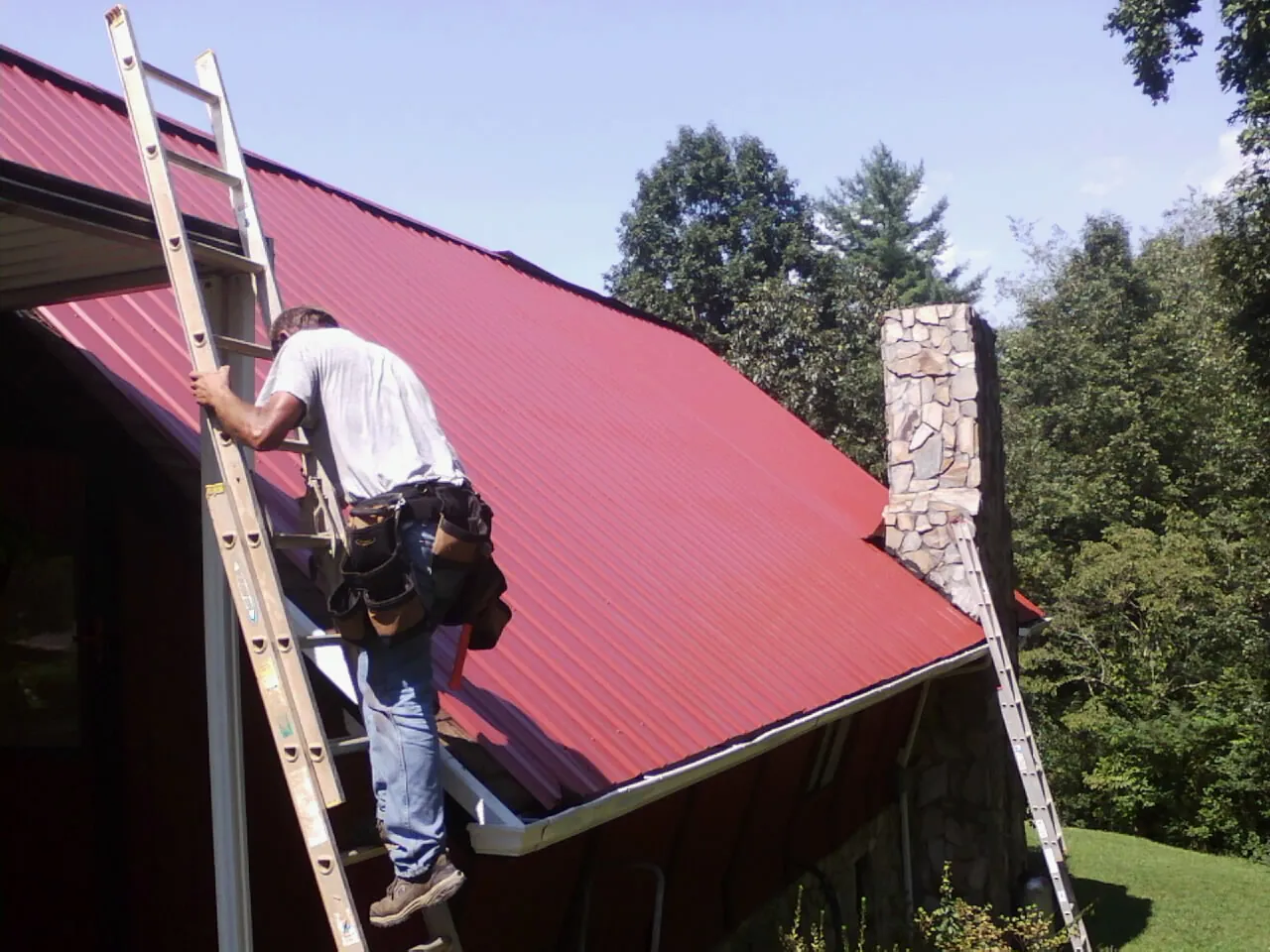 Roofer on a ladder beside a red metal roof and stone chimney — Soffit & Fascia Repair by Roofing Brothers in Olivebridge